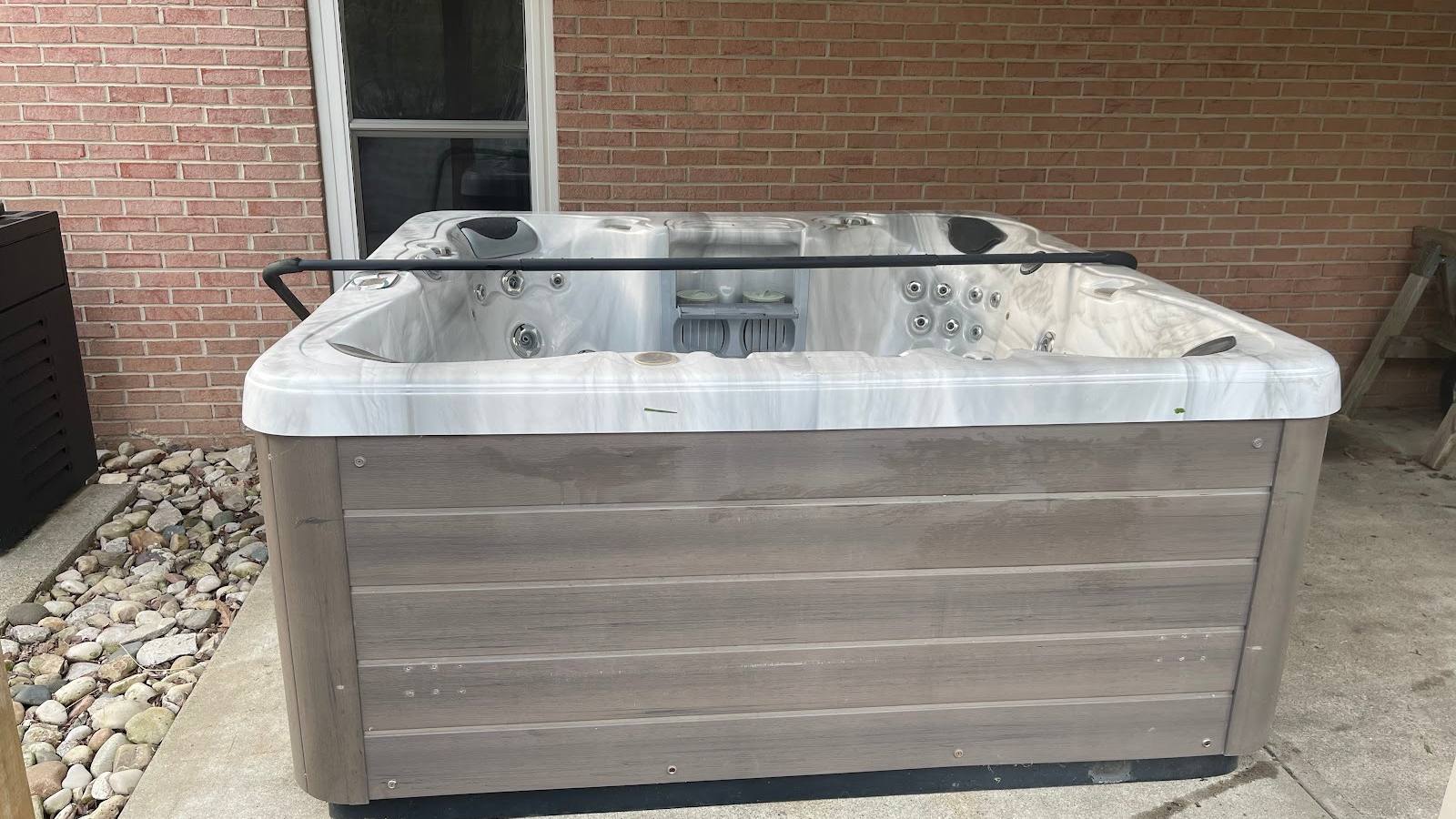 A large light grey marbled acrylic hot tub with its cover open, showing the moulded seats and jets, on a concrete patio beside a brick wall.