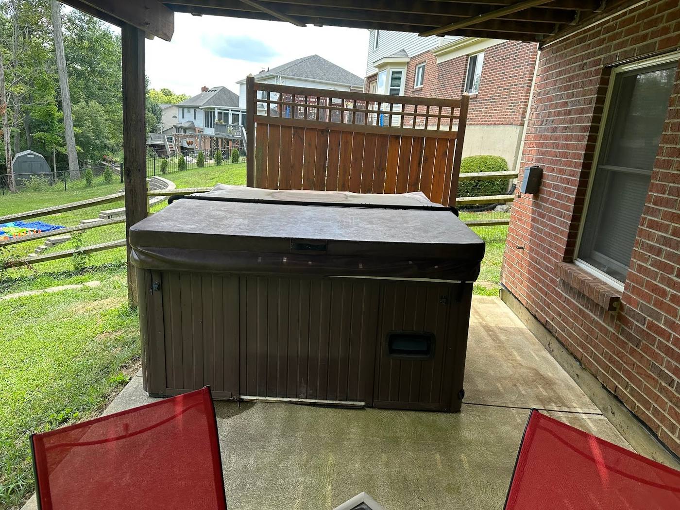 A dark brown hot tub with a grey cover standing on a concrete patio tucked under the overhang of a brick house, with a wooden privacy screen behind it.