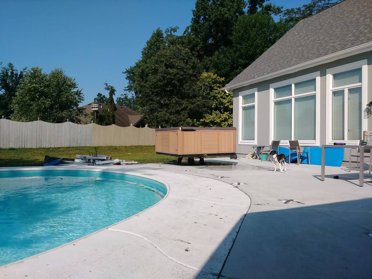 A wood-sided hot tub standing on the concrete deck of a kidney-shaped in-ground pool behind a house, with a privacy fence and trees beyond.