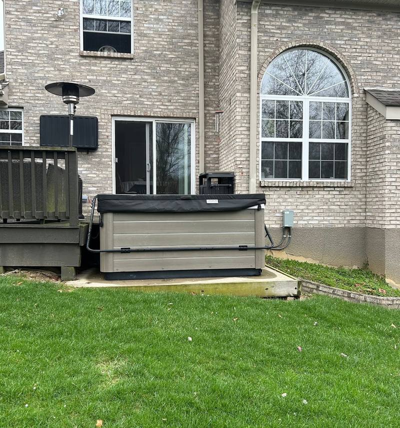 A grey hot tub with a dark cover on a concrete pad beside a brick house, next to a wooden deck, with a green lawn in front.