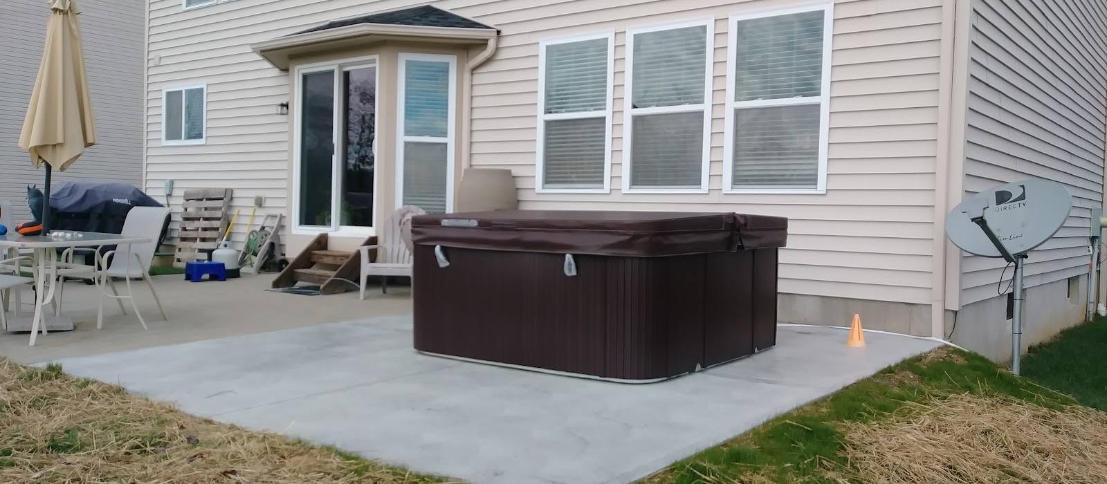 A dark brown hot tub on a wide new concrete patio behind a cream-sided house, with patio furniture and a folded umbrella nearby.